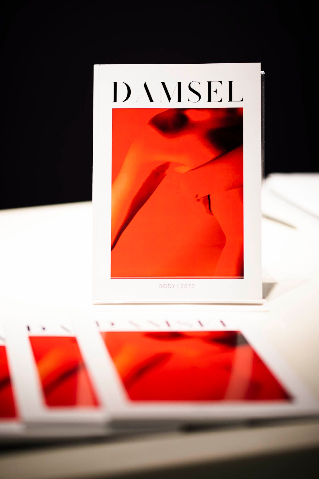 Copies of Damsel Magazine are displayed on a stark white tablecloth. The magazine is glossy, with a dramatic contrast between the abstract red photograph of a person's torso and the crisp black-and-white title. Photograph by Charlie Beveridge, courtesy of the UWA Student Guild Women's Department.